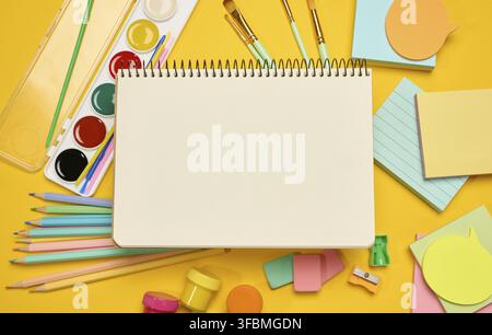 Open blank spiral notebook in the center, surrounded by school supplies colored pencils, watercolors, brushes, erasers and sticky notes, yellow backgr Stock Photo