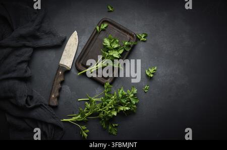 Green sprigs of parsley on a wooden cutting board and a knife. Top view of black kitchen table Stock Photo