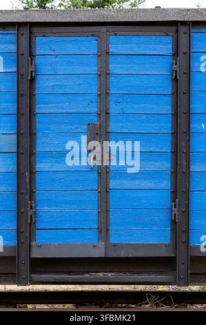 Railway wagon side with a closed blue wooden door. Padlock secures a metal latch on the door. Rivets and bolts fasten the frame and wooden planks. Stock Photo