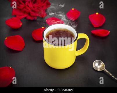 Yellow cup with hot coffee on a black background among the petals of a red rose Stock Photo