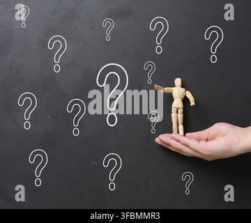 A woman's hand holding a wooden miniature puppet against a black chalkboard with chalk-drawn question marks. Concept of searching for truth, gathering Stock Photo