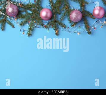 Burning multi-colored light bulbs on a garland with a white cable and green twigs, pink balls, blue background, copy space Stock Photo