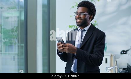 Happy cheerful African American business man smiling scrolling mobile phone app in office carefree businessman male guy executive agent using Stock Photo