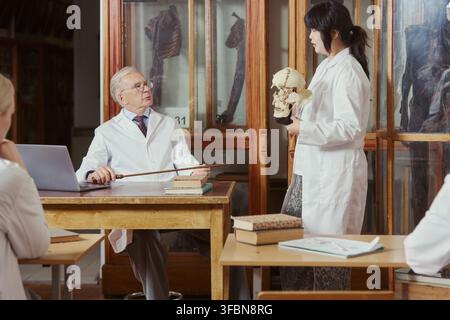 Asian female young adult student holding skull model, presenting to senior Caucasian male professor in anatomy lab. Image suitable for medical university recruiting and higher education marketing Stock Photo