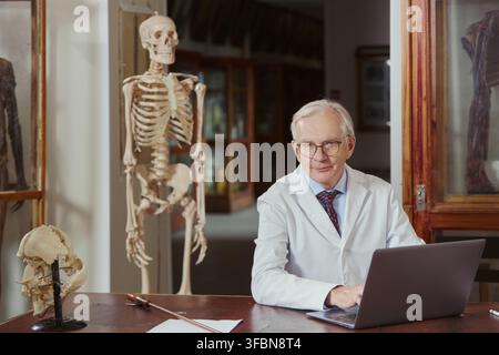 Portrait of senior Caucasian male scientist using laptop in anatomy museum. Image represents medical education and research, suitable for university brochures and healthcare marketing Stock Photo