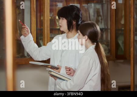 Asian young adult and Caucasian young adult female students wearing lab coats, studying museum exhibits and taking notes. Image suitable for university education and science research marketing Stock Photo