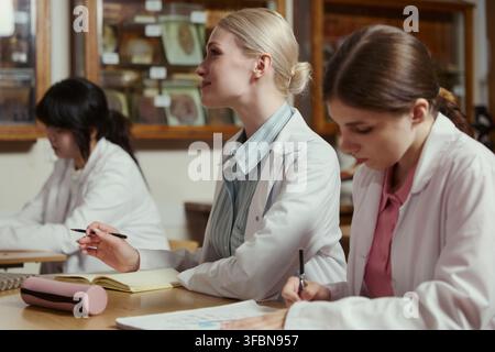 Diverse female young adult students in lab coats taking notes during university science lecture. Image represents higher education and is suitable for academic marketing materials Stock Photo
