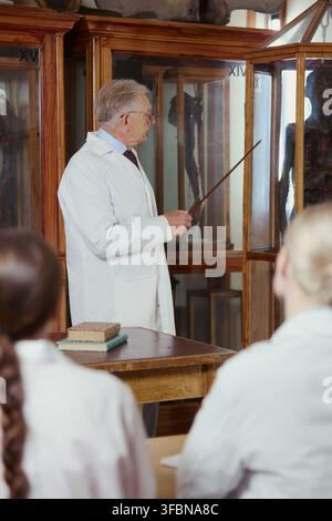 Senior Caucasian male professor in lab coat pointing at anatomical model during lecture for medical students. Concept of higher education and anatomy training, vertical Stock Photo