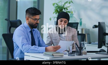 Two concentrated Indian business colleagues discussing papers graphs financial data results talking working computer in office. Focused Arabian man Stock Photo