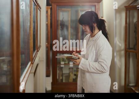 Asian young adult female researcher wearing lab coat writing in notebook while examining museum exhibits. Image suitable for academic research and science education marketing Stock Photo