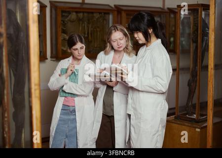 Diverse young adult female medical students in lab coats taking notes in anatomy museum. Image suitable for university brochures and medical education marketing Stock Photo