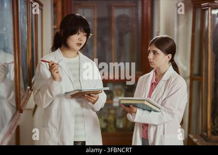 Asian young adult and Caucasian young adult female students wearing lab coats, holding notebooks and discussing research in museum. Image suitable for university education marketing Stock Photo