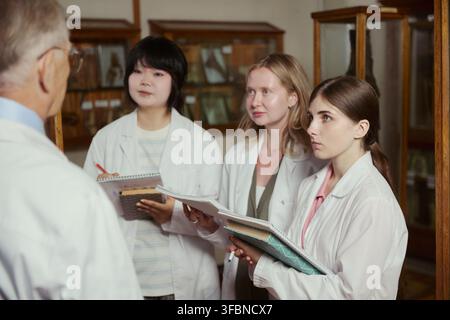 Diverse group of young adult female medical students in lab coats listening to senior male professor in museum. Image suitable for university brochures and medical education marketing Stock Photo