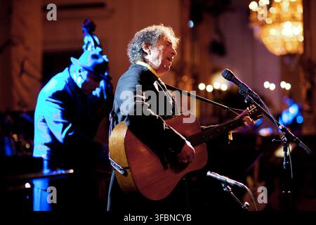 Musician Bob Dylan performs on stage, playing an acoustic guitar and singing into a microphone, during a concert in 2010. He is seen from the side, with curly hair, wearing a dark suit jacket, against a blurred background with warm lighting and a chandelier. Another musician, illuminated by blue light, is partially visible in the background. Pete Souza photo (White House photo) Stock Photo