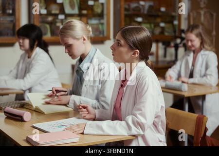 Diverse group of female Caucasian and Asian teenagers wearing lab coats, sitting at desks and taking notes during biology class. Image ideal for high school education marketing Stock Photo