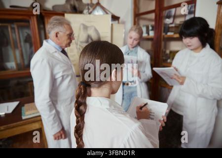 Diverse young adult medical students in lab coats taking notes during lecture with senior Caucasian male professor in anatomy classroom, perfect for medical education marketing Stock Photo
