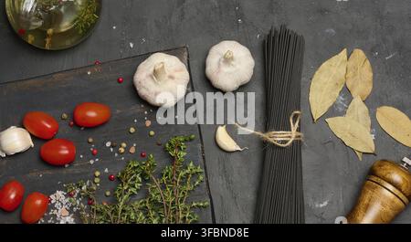 A bunch of black spaghetti, a wooden mill, heads of garlic, a bunch of thyme and a bay leaf on a dark textured background Stock Photo