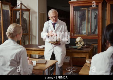 Senior Caucasian male professor holding book and lecturing young adult students in anatomy museum. Image suitable for medical education and university recruitment campaigns Stock Photo