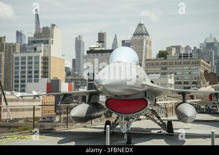 Front view of a modern gray fighter jet with red intake cover, displayed at an outdoor aviation museum. City skyscrapers form a dramatic backdrop behi Stock Photo