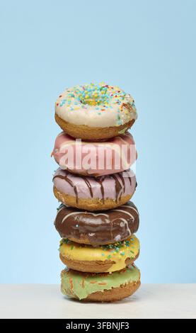 A tall stack of six colorful, glazed donuts with various toppings is centrally placed on a white surface against a solid light blue background Stock Photo