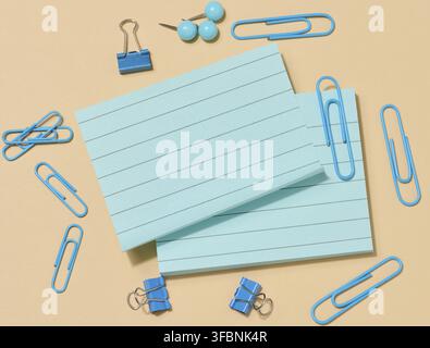 Several stacks of light blue sticky notes and many blue paper clips of various shapes, arranged on a beige background. Top view Stock Photo