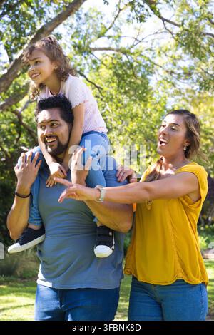 Diverse family with child walking in sunlit park under leafy canopy with man carrying girl Stock Photo