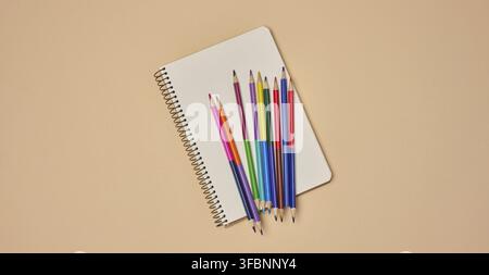 Top view of an open notebook with pastel-colored pencils on a beige background. Flat lay composition of school or office stationery supplies Stock Photo