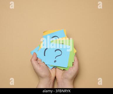 Female hands hold paper sheets with a drawn question mark on a beige background, searching for the answer and truth Stock Photo