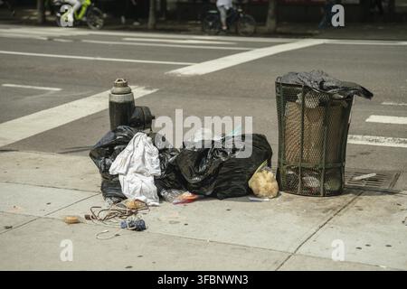 Black garbage bags and loose trash lie next to an overflowing street bin on a concrete sidewalk. A fire hydrant and urban crosswalk mark the busy city Stock Photo