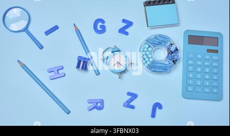 Flat lay of blue stationery, learning items letters, magnifying glass, supplies, and an alarm clock on a blue background. Education, time, and study c Stock Photo