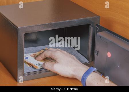Hand taking stack of euro banknotes from open small black metal safe box on wooden shelf in hotel room stock photo. Stock Photo