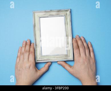 Two female hands holding a rectangular blank wooden frame, blue background Stock Photo