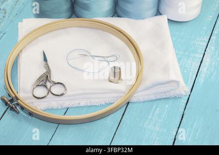Wooden round embroidery frame and white fabric for cross stitching, behind a row of spools with threads Stock Photo