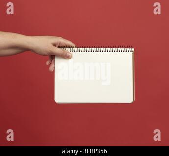 Hand holding an open spiral notebook with empty white sheets, red background, copy space Stock Photo