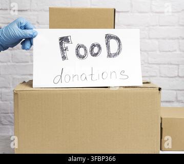 Stack of cardboard boxes and white sheet of paper with inscription food donation on white brick wall background Stock Photo