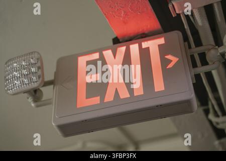 Close-up of a red EXIT sign with built-in emergency light, mounted on a ceiling near electrical conduit. Symbol of safety and emergency evacuation Stock Photo