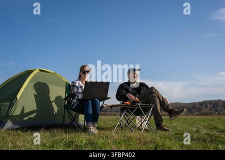 Couple camping in a sunny meadow: woman focused on laptop working remotely while man relaxes by the tent, enjoying nature, freedom and a peaceful outd Stock Photo