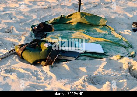 Open book lies on green military blanket on sandy beach. Warm sunlight highlights sand and fabric textures. Scene suggests peaceful reading during sea Stock Photo