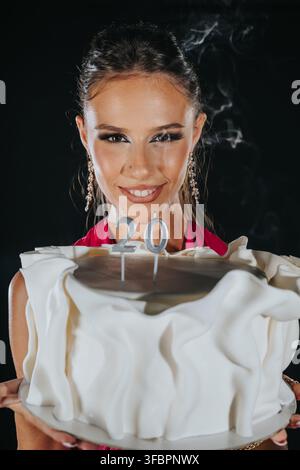 A young woman holds a white-frosted birthday cake topped with number 20 candles. She smiles while presenting the cake in a celebratory party pose. Stock Photo