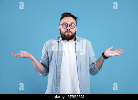 Who knows. Confused bearded man in glasses shrugging shoulders on blue studio background Stock Photo