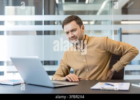 Businessman sitting at a desk and working on a laptop, experiencing discomfort and pain in his lower back, highlighting issues with prolonged sitting and poor posture Stock Photo