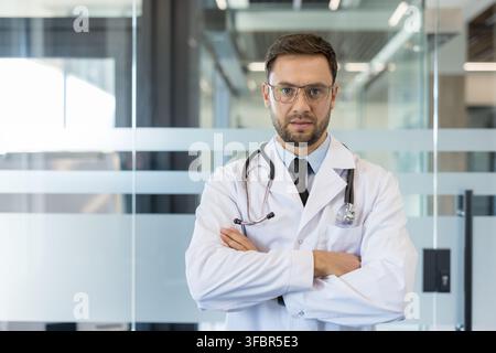 Professional male doctor wearing a white lab coat and stethoscope, standing with arms crossed, conveying expertise and confidence in a modern healthcare environment Stock Photo