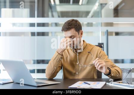Stressed businessman feeling tired and suffering from a headache, rubbing his nose bridge while holding eyeglasses, experiencing burnout from work at his modern office desk Stock Photo