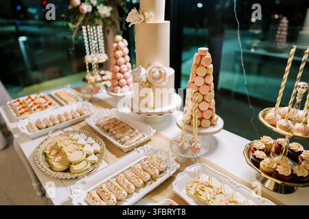 elegant wedding cake displayed on a dessert table with macarons and cupcakes in front-facing, slightly elevated angle with pink and white colors and Stock Photo