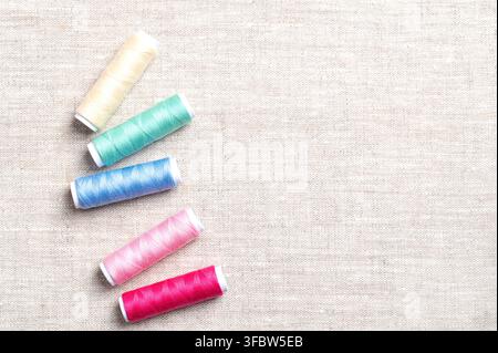 Polyester sewing thread spools randomly distributed on linen fabric, from above, with copy space. Multi colored vibrant polyester sewing thread. Stock Photo