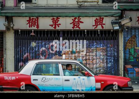 A red and white Hong Kong taxi, with a driver visible inside, moves across the frame past a closed storefront featuring a metal rolling shutter with b Stock Photo