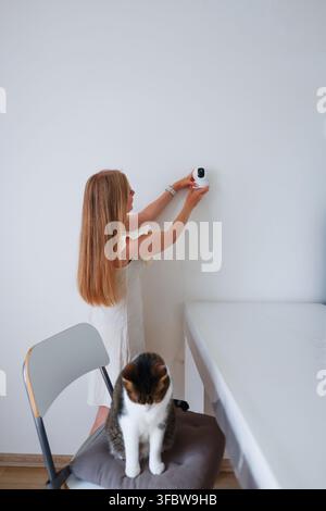 Woman mounting an indoor surveillance camera on the wall with a curious cat nearby, smart home security concept. Stock Photo