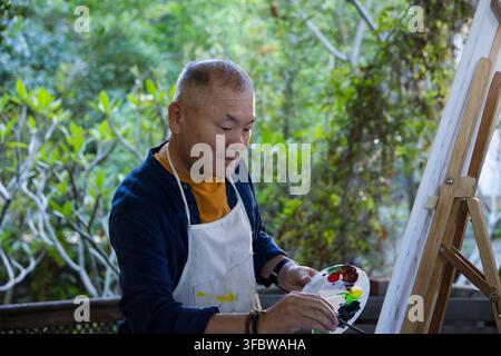 Senior Asian man in paint-stained apron, standing on porch at wooden easel mixing paint on palette. Elder, painter, canvas, patio, garden, outdoor, fo Stock Photo