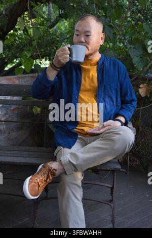 Senior Asian man sitting on wooden bench on deck sipping from grey ceramic mug wearing wristwatch Stock Photo