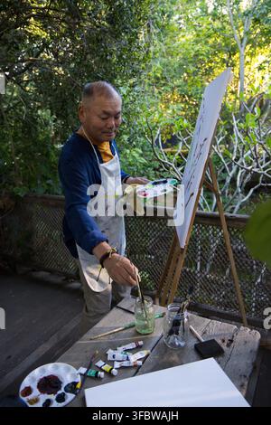Senior Asian man painting at easel on deck, holding palette and brushes, wearing painted apron Stock Photo